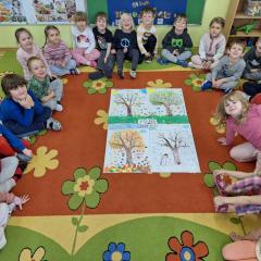 dzieci siedzą na dywanie i prezentują swoje prace grupowe