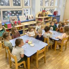 Dzieci siedzą przy stolikach i słuchają instrukcji tworzenia świeczek